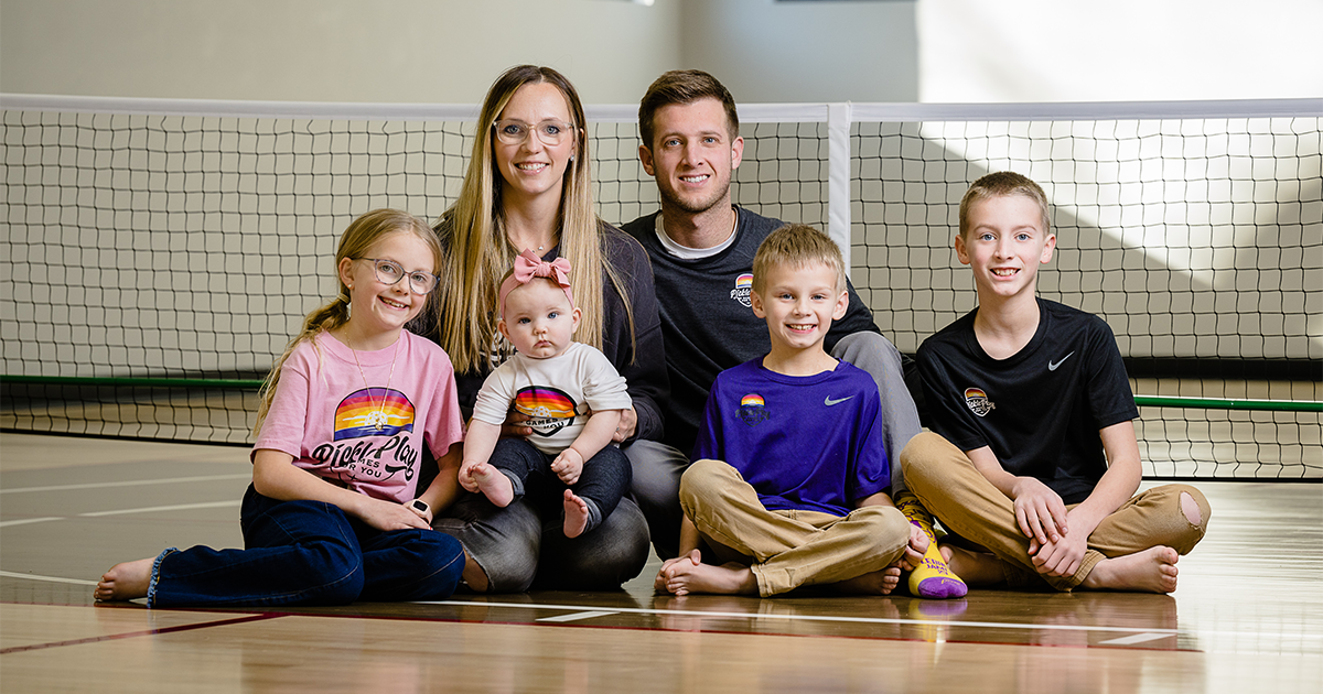 Blake, her husband, and their four children sit on an indoor pickleball court, smiling at the camera.