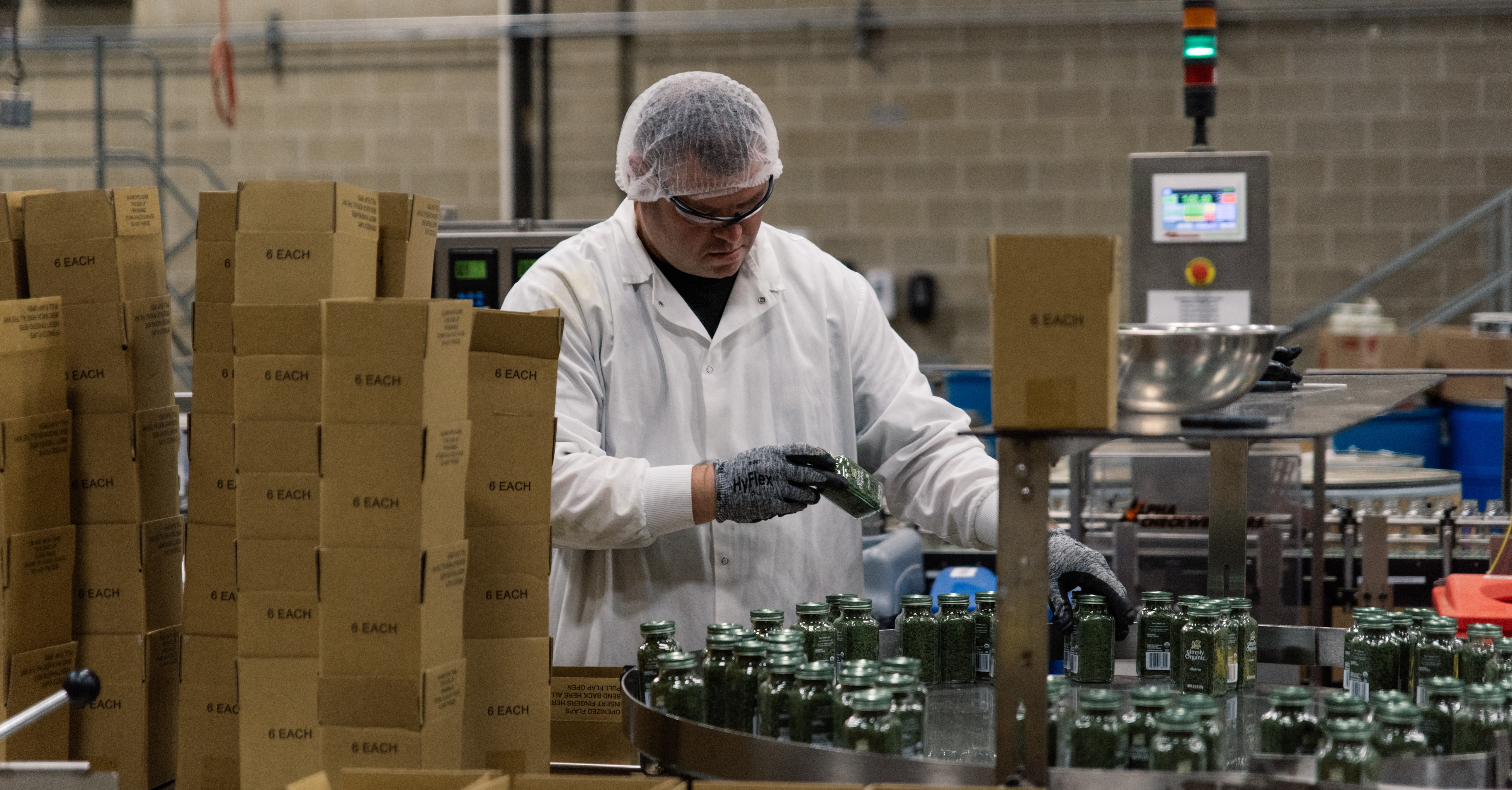 A man wearing a hair net and white lab coat checks spice jars for quality.