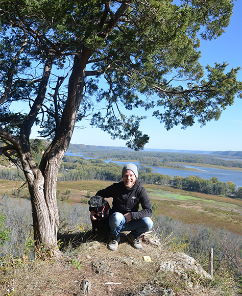 Hiker and his dog smile at the camera from atop a riverside hill. 