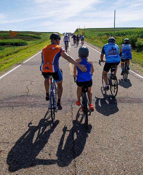 A father and daughter bike on a road passing corn fields with other cyclists