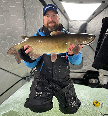 Man kneeling in his ice hut holding up a large fish.