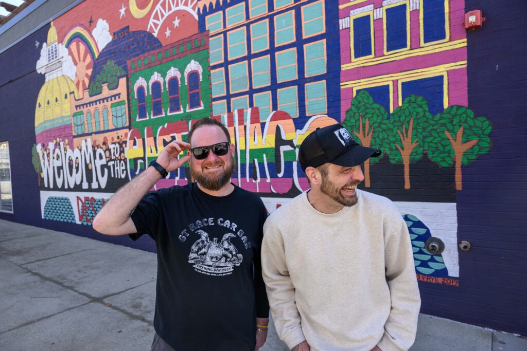 Phil Shires and Nick Hanke stand laughing in front of a brightly-colored mural that reads, "Welcome to the East Village."