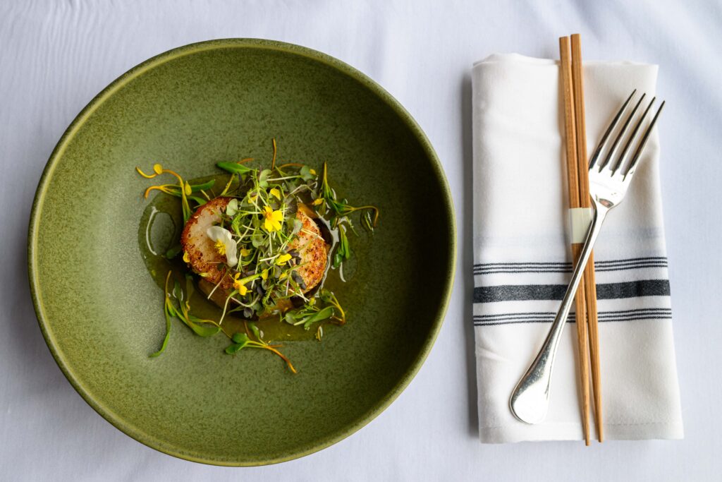 A green plate sits to the right of a fork and chopsticks on top of a white napkin and white table cloth.