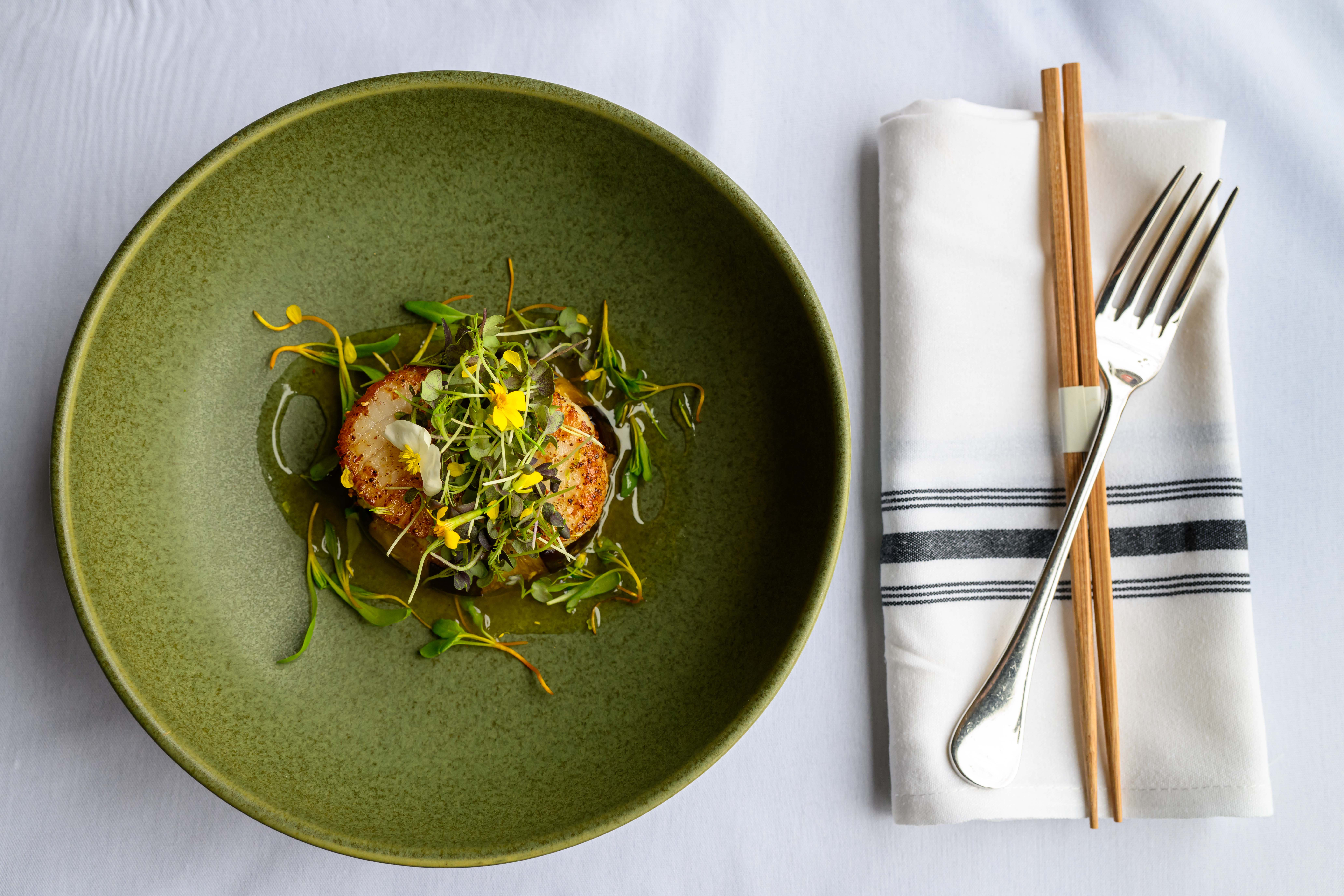 A green plate sits to the right of a fork and chopsticks on top of a white napkin and white table cloth.