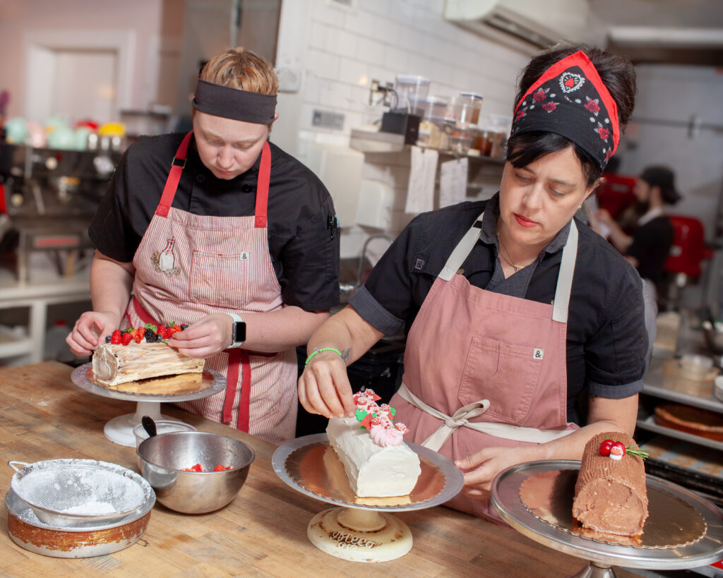 Two women, including baker Jamie Powers, work in a kitchen setting. They are standing at a wooden table while working on three log style cakes