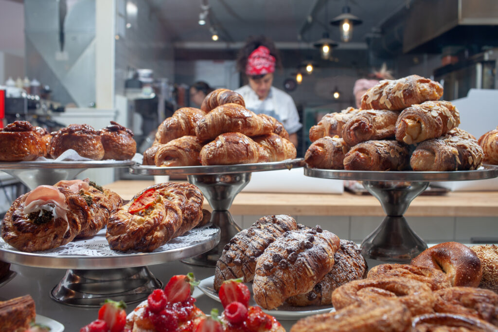 Assorted pastries, including croissants and Danishes, are displayed on metal stands in a bakery with staff working in the background