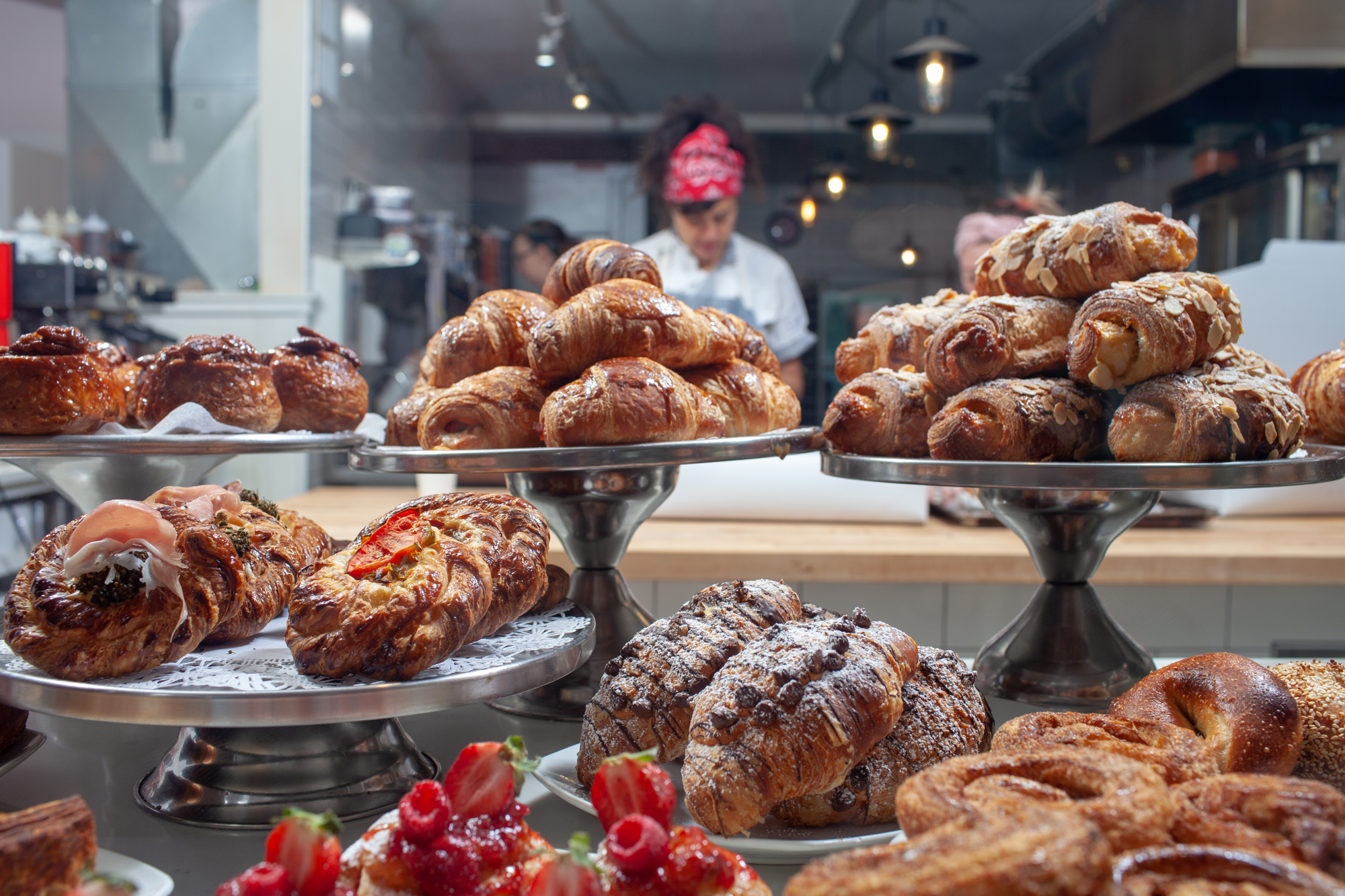 Assorted pastries, including croissants and Danishes, are displayed on metal stands in a bakery with staff working in the background