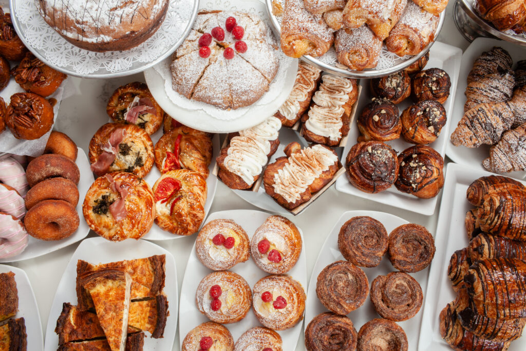An assortment of pastries and baked goods from DeLuxe bakery on while platters, featuring items like a powdered cake with raspberries, croissants, Danishes, and cinnamon rolls