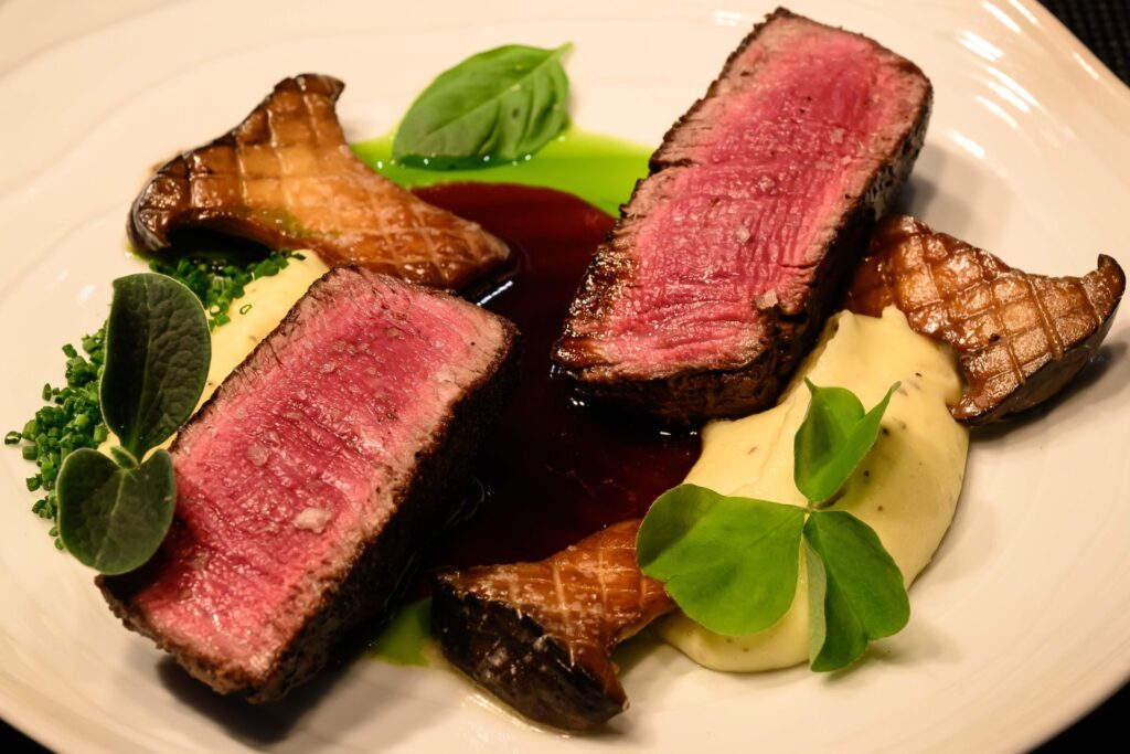 A plated gourmet dish featuring two slices of medium-rare steak, seared mushrooms, and dollops of creamy mashed potatoes, garnished with fresh green herbs. The dish is presented on a white plate with contrasting colors of pink meat, brown mushrooms, green leaves, and light yellow mashed potatoes, highlighting a sophisticated culinary presentation.