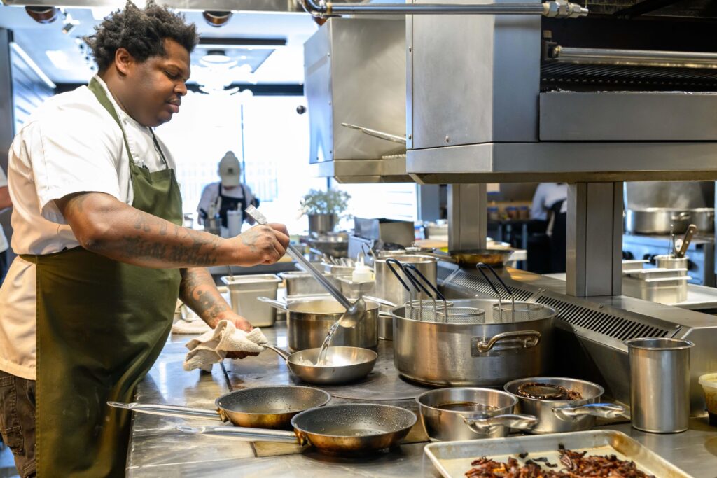 Chef Ian Robertson stands in a professional kitchen ladling water into a pan, surrounded by various pots, pans, and cooking utensils on a stainless steel counter. The scene highlights a busy cooking environment with multiple dishes being prepared simultaneously, emphasizing culinary activity and kitchen organization.
