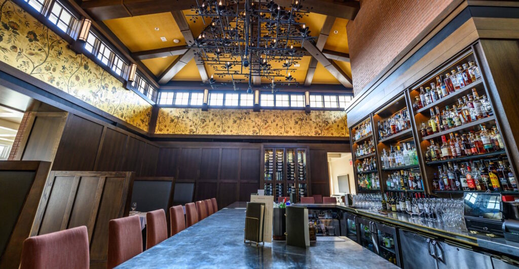 An upscale bar interior featuring a long marble countertop with pink cushioned chairs and a large wooden shelving unit stocked with various liquor bottles. The space has high ceilings with exposed wooden beams, decorative floral wallpaper, and a distinctive black metal chandelier with leaf motifs.